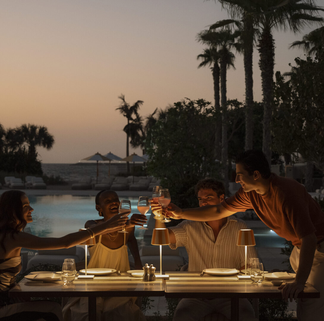 Friends toast drinks at dusk by a pool, Banyan Tree Dubai-Tutto Passa restaurant with palm trees in the background.
